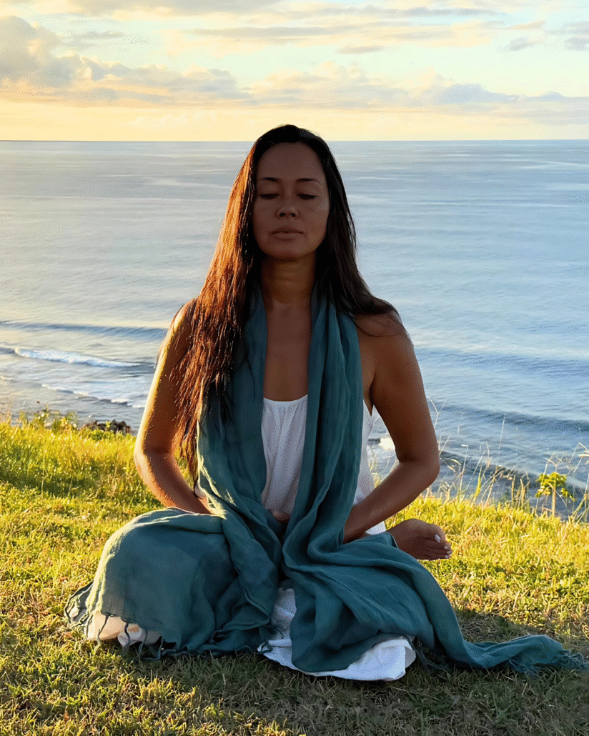 Dr. Satya Shiva meditating in lotus pose on the ocean cliffs of Princeville, Kauai, with waves below and blue sky above.